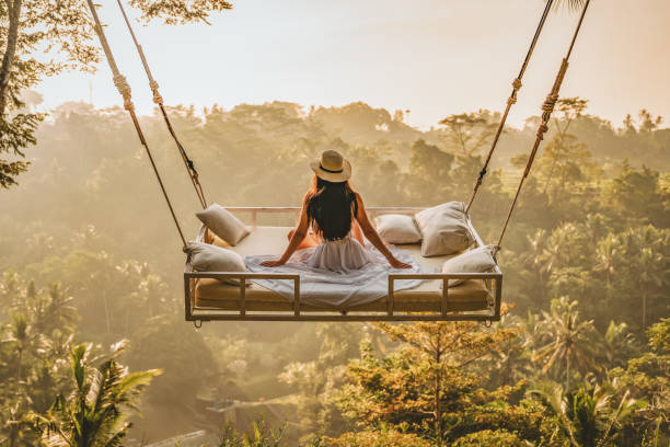 Photo of a young woman sitting on the swing. Jungle Bed hanging over the tropical forest with Caucasian female resting while looking at the view, Bali, Indonesia. Rear view of a female sitting and enjoying the view.