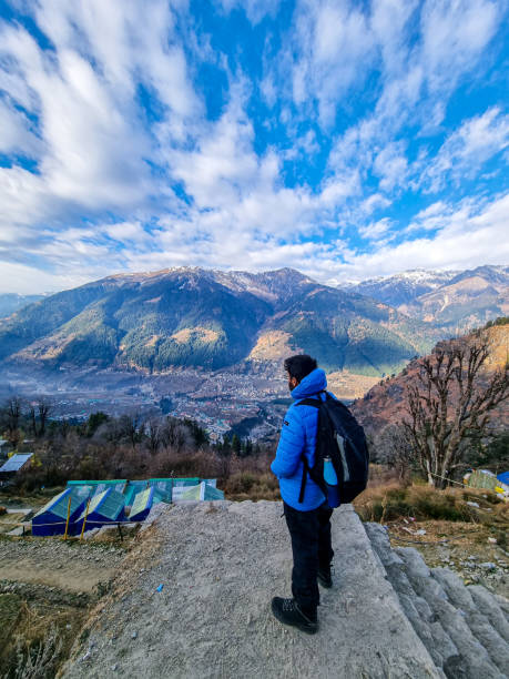 Male trekker hiker standing looking down at village houses and himalaya mountains in distance and cloudy sky in manali, kullu, shimla, kedarnath in India