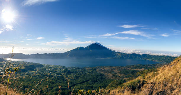 A breathtaking view showcasing Mount Batur in Bali, Indonesia, during a serene morning, captured with lush greenery, a calm lake, and clear blue skies for an enchanting natural landscape scenery.
