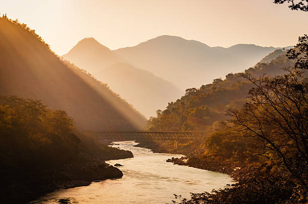 A mesmerizing view of the serene sunrise over the Ganga River in Rishikesh, India, with a historic bridge in the background. The golden morning light reflects off the holy river, creating a peaceful and spiritual atmosphere surrounded by the stunning Himalayan foothills